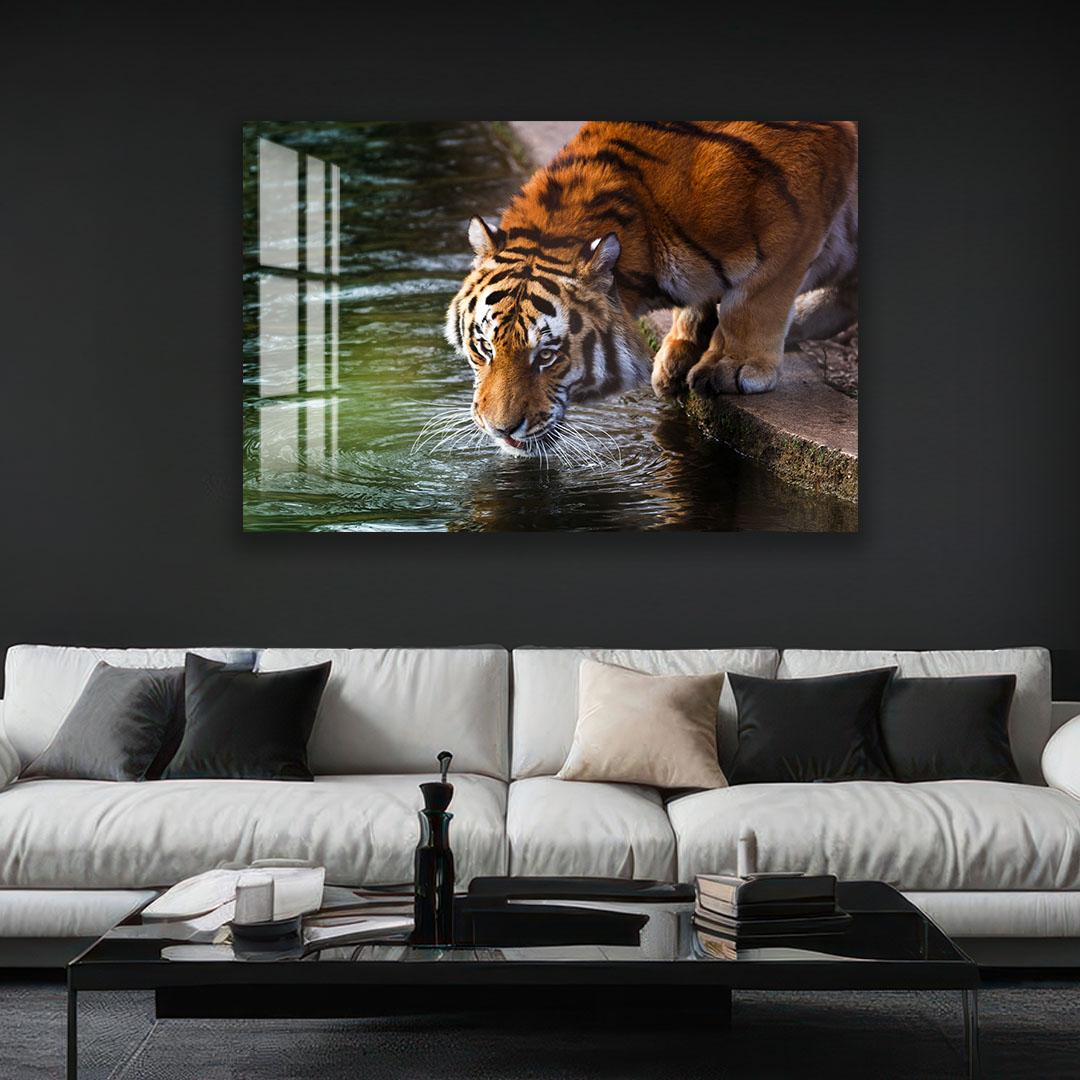 A tiger is drinking from a pond in a lush green forest, as depicted in a large framed artwork hanging on a dark wall above a white sofa in a modern living room setting.