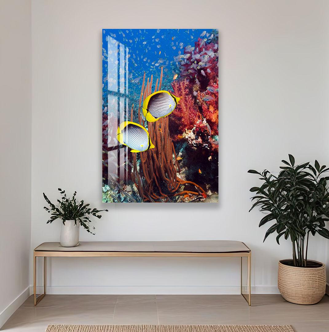 a large painting of a vibrant underwater scene with two fish swimming among coral and plants. The painting is hung on a white wall above a wooden bench, which is positioned in front of a potted plant.