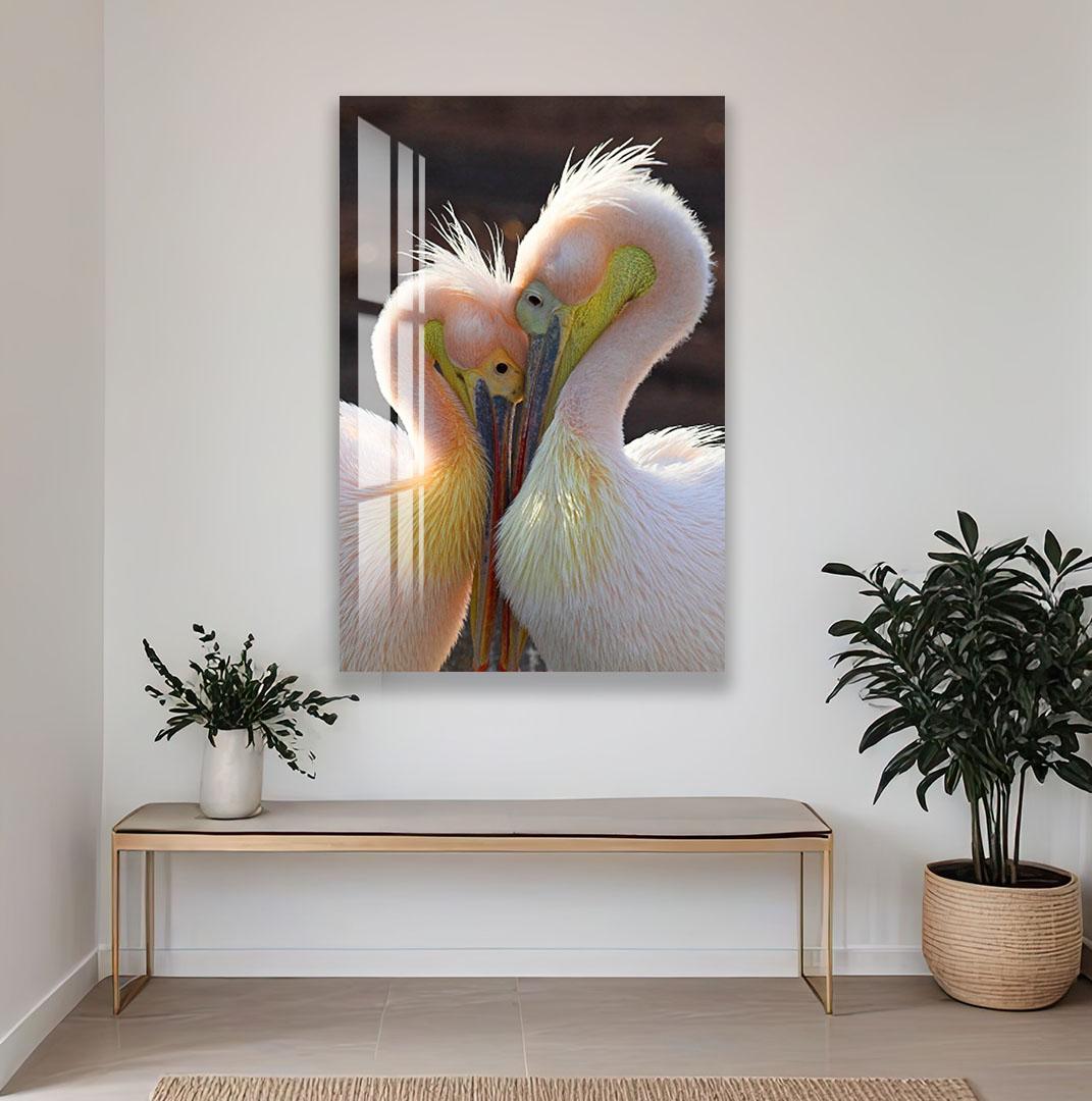 a large, colorful painting of two pelicans with their heads close together, creating an intimate and affectionate moment. The painting is hung on a white wall above a wooden bench, which is positioned in front of a potted plant. The overall scene is set in a room with a rug on the floor, adding to the cozy and inviting atmosphere.