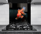 A vibrant orange goldfish is captured in mid-swim, its fins spread wide, against a backdrop of a black stovetop and white tiled wall.